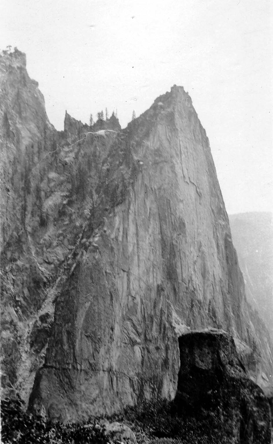 A towering, sheer granite formation rising sharply from forested talus slopes into the sky in Yosemite Valley.