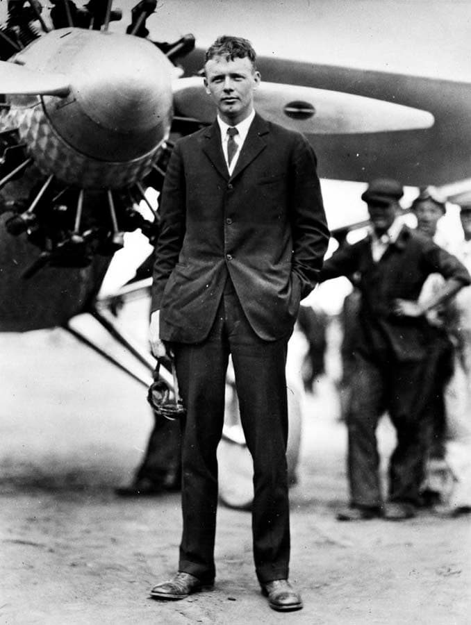 Full-length portrait of Charles A. Lindbergh in dark suit holding goggles in front of the Spirit of St. Louis at Dutch Flats.