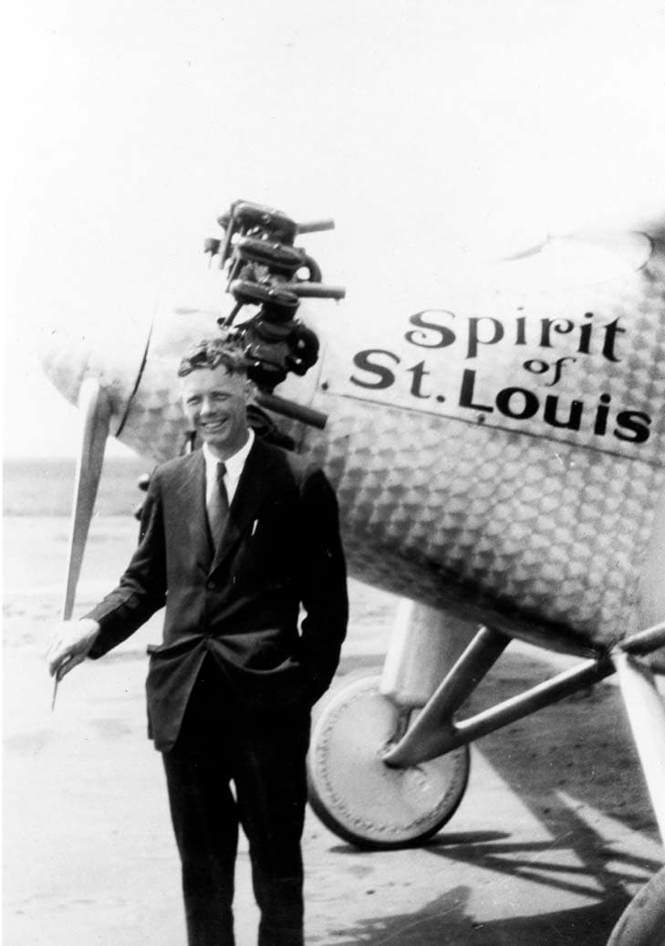Close-up photograph of Charles Lindbergh smiling beside the damascened nose cowl and painted Spirit of St. Louis insignia.