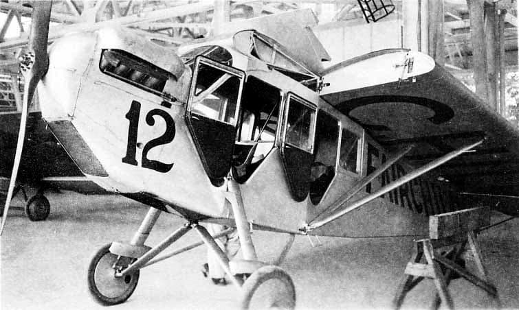 A high-wing commercial passenger monoplane marked number 12 parked inside a hangar with three cabin doors standing open.