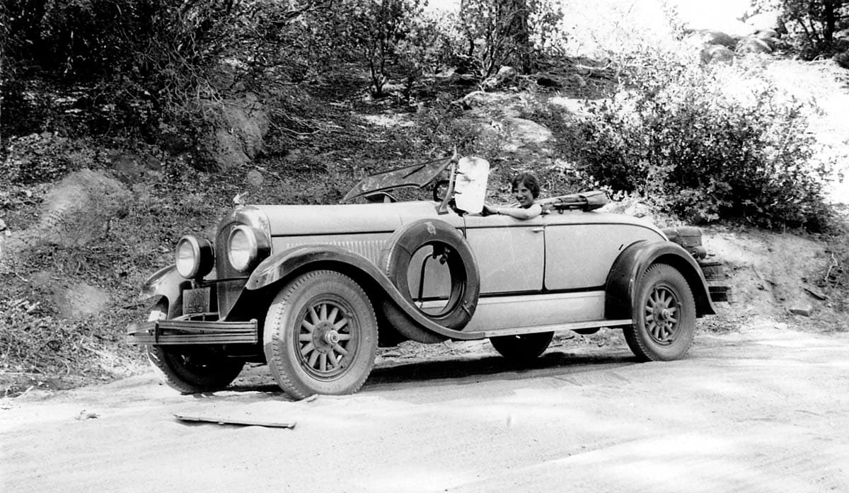 A smiling woman seated at the wheel of Donald A. Hall Sr.'s 1925 Studebaker Roadster on a rugged dirt mountain road.
