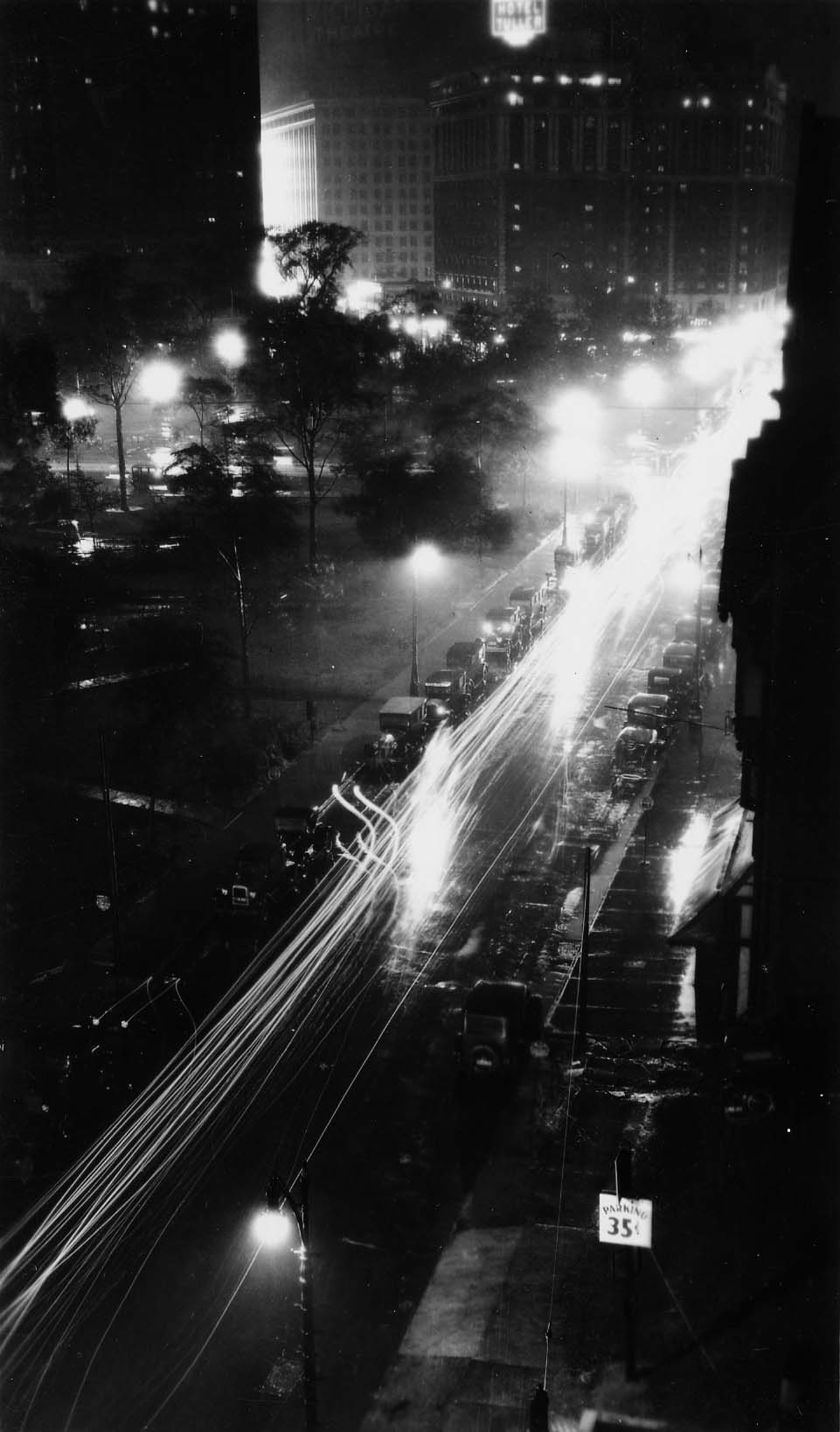 A long-exposure night streetscape from above, capturing streaking automobile headlights along a wet street leading to the illuminated Hotel Tuller.