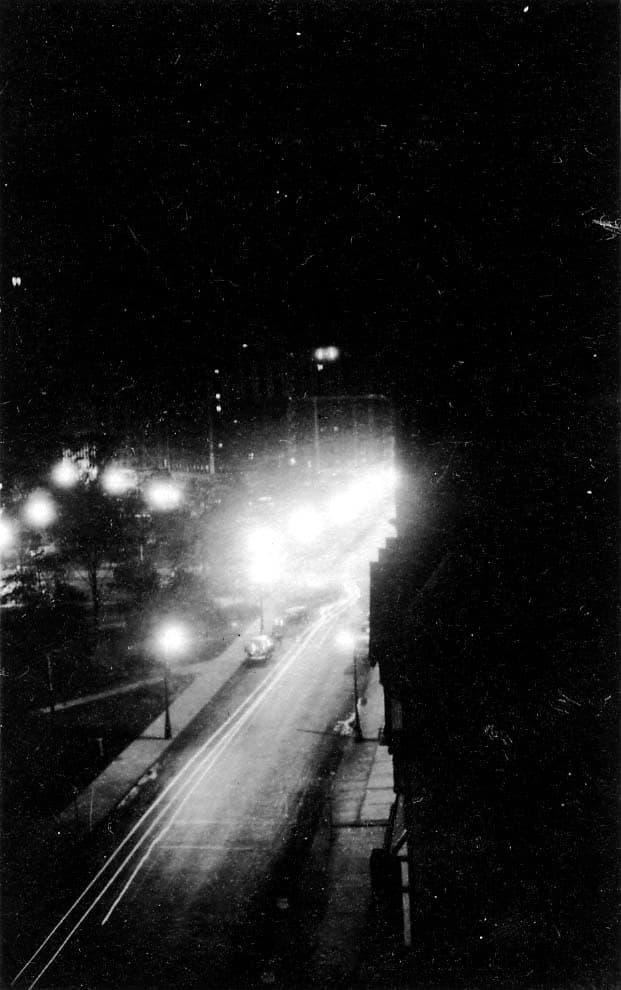 A high-angle vertical night perspective down a brightly lit Detroit avenue flanking Grand Circus Park, showing streaks of moving traffic beneath rows of glowing streetlights.