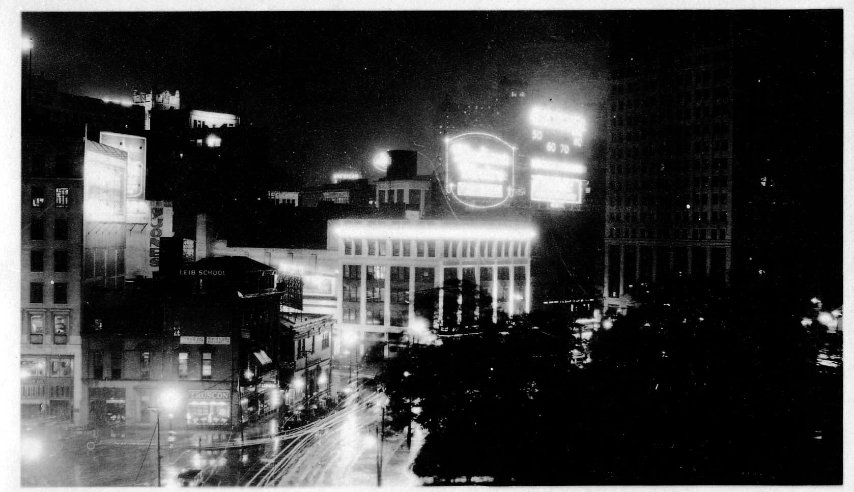 A wide-angle night photograph across Grand Circus Park toward the glowing Madison Theatre sign, illuminated commercial blocks, and the David Whitney Building under a dark sky.
