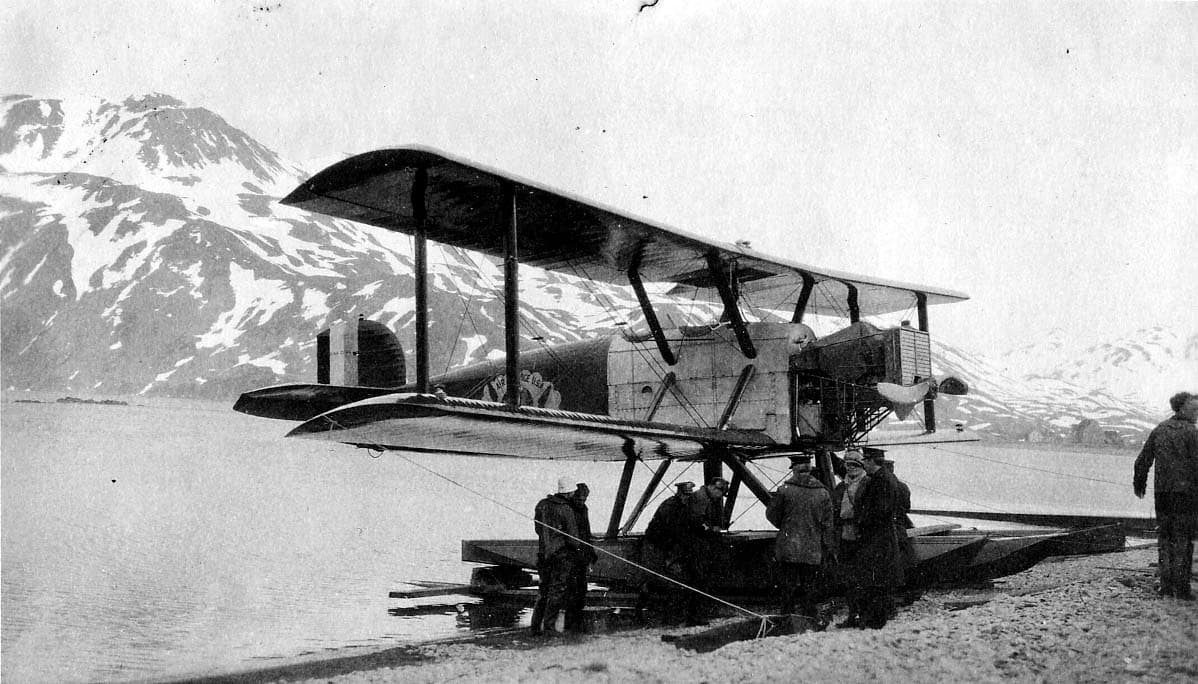 A Douglas World Cruiser seaplane beached on shingle shoreline with military crew in winter coats gathered under the wing.