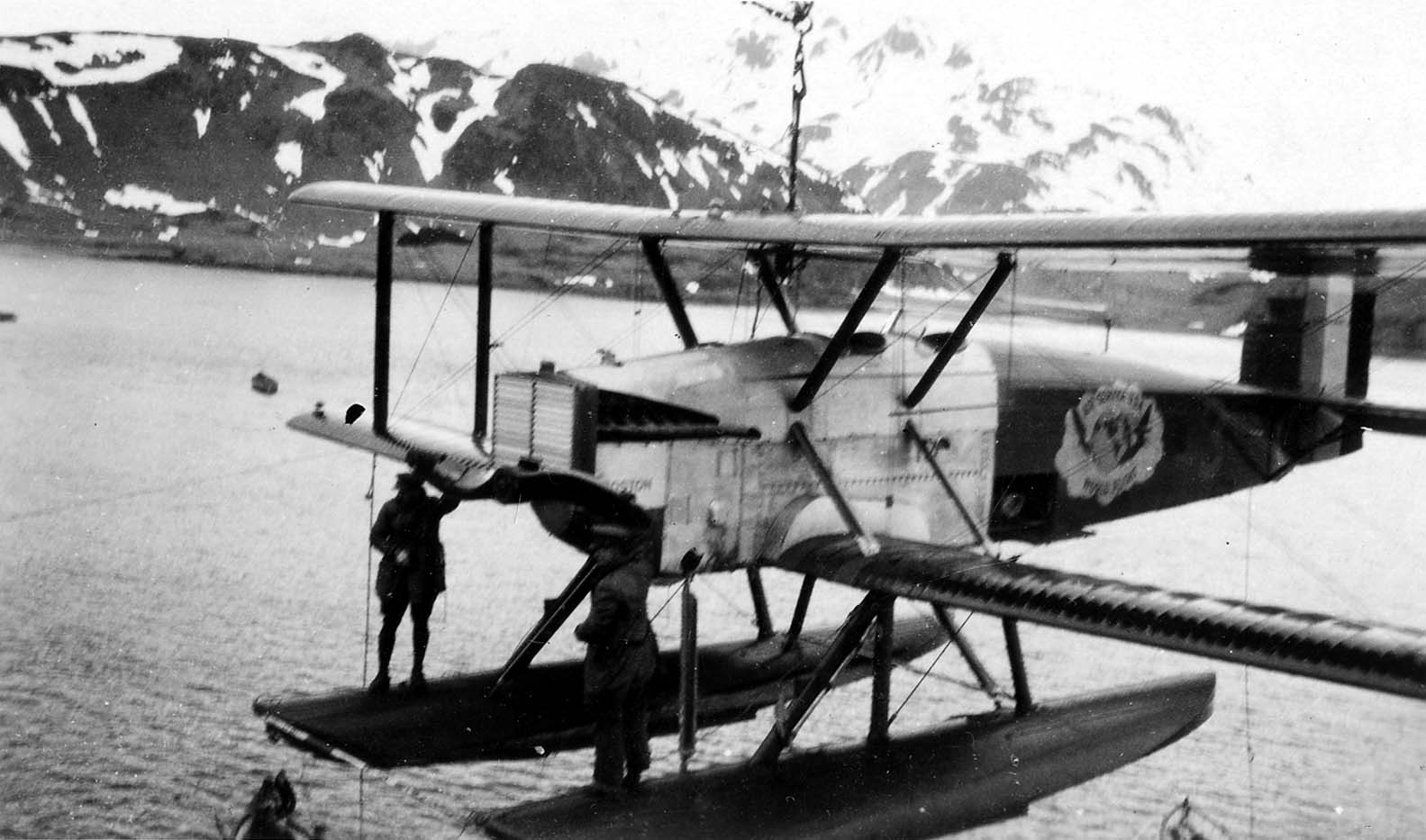 The Douglas World Cruiser Boston with floats suspended in mid-air by a ship's crane against snow-capped Alaskan mountains.