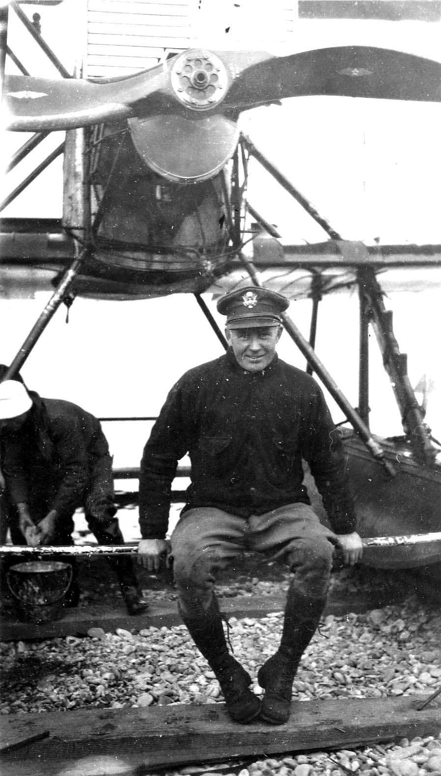 A smiling aviator in an Army Air Service cap and cold-weather flight gear seated on the pontoon spreader bar beneath the massive propeller of a Douglas World Cruiser seaplane.