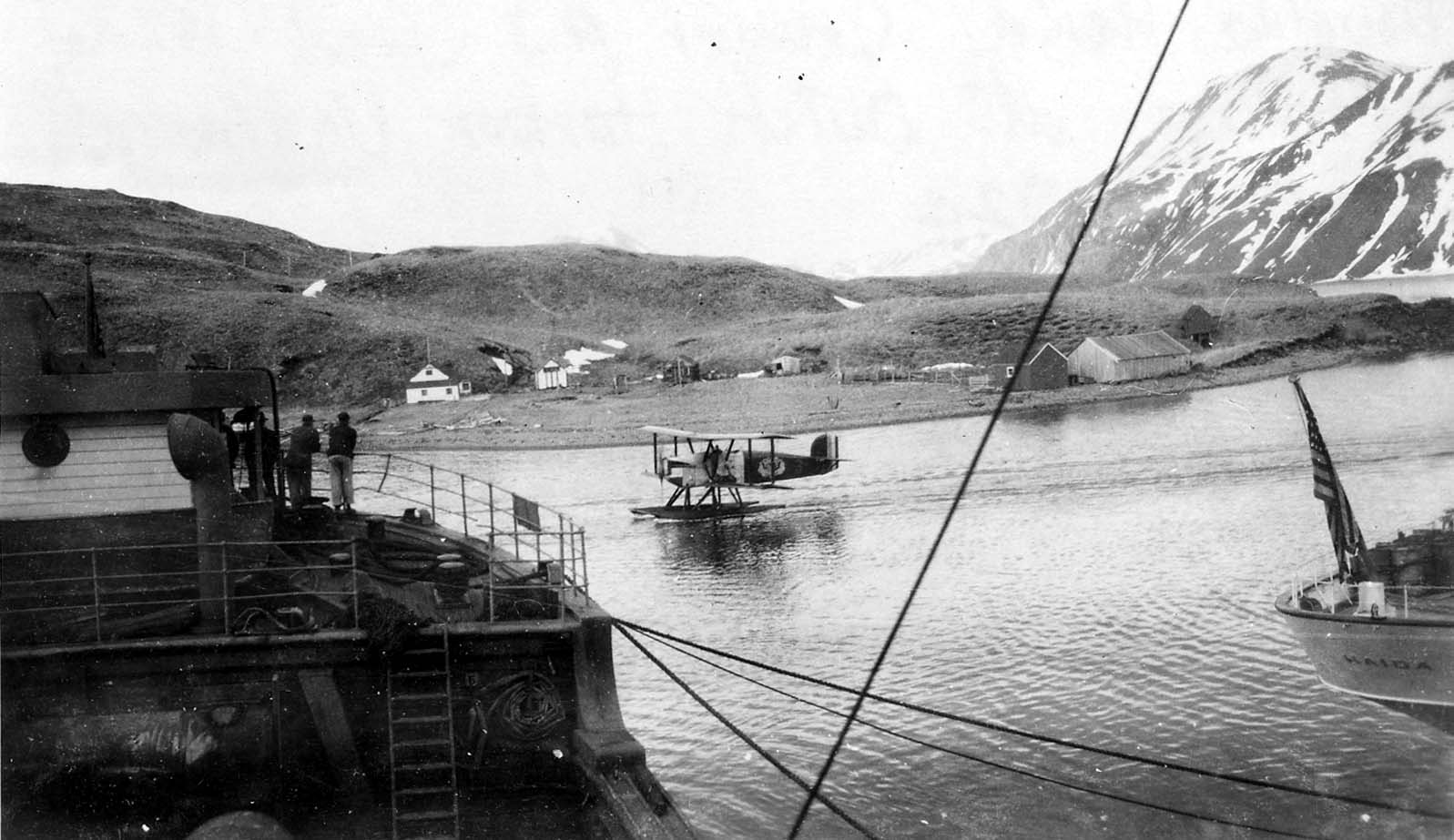 A Douglas World Cruiser seaplane taxiing in a bay near the stern of the U.S. Coast Guard Cutter Haida flying an American flag.