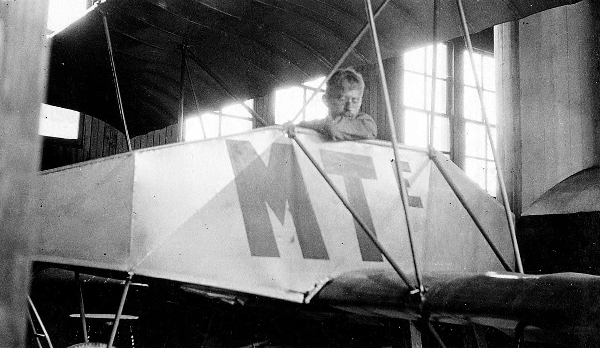 A pilot seated in the cockpit of an experimental light monoplane hand-lettered MTE inside a wooden hangar workshop.