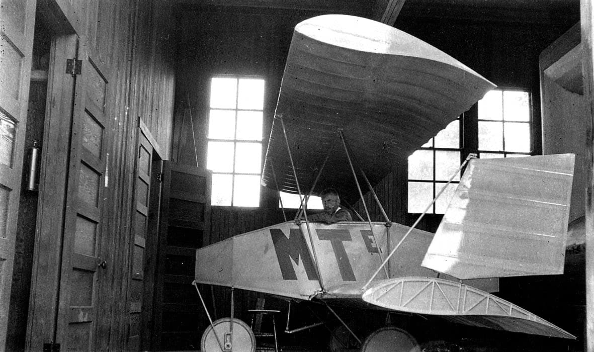 A rear three-quarter view of the experimental MTE light monoplane inside a wooden workshop showing its large rectangular tail and wing ribs.