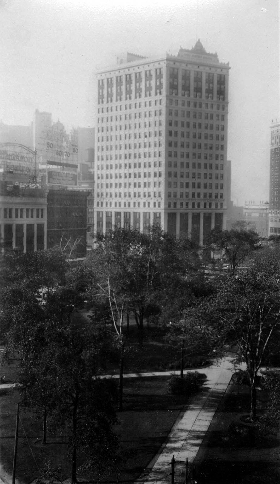 A high-angle view looking down through the trees and walkways of Grand Circus Park toward the towering David Whitney Building.