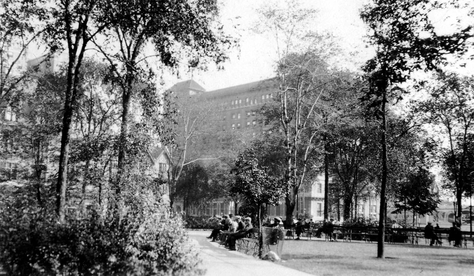 Pedestrians resting on wooden benches beneath leafy shade trees in Grand Circus Park with downtown Detroit commercial buildings behind.