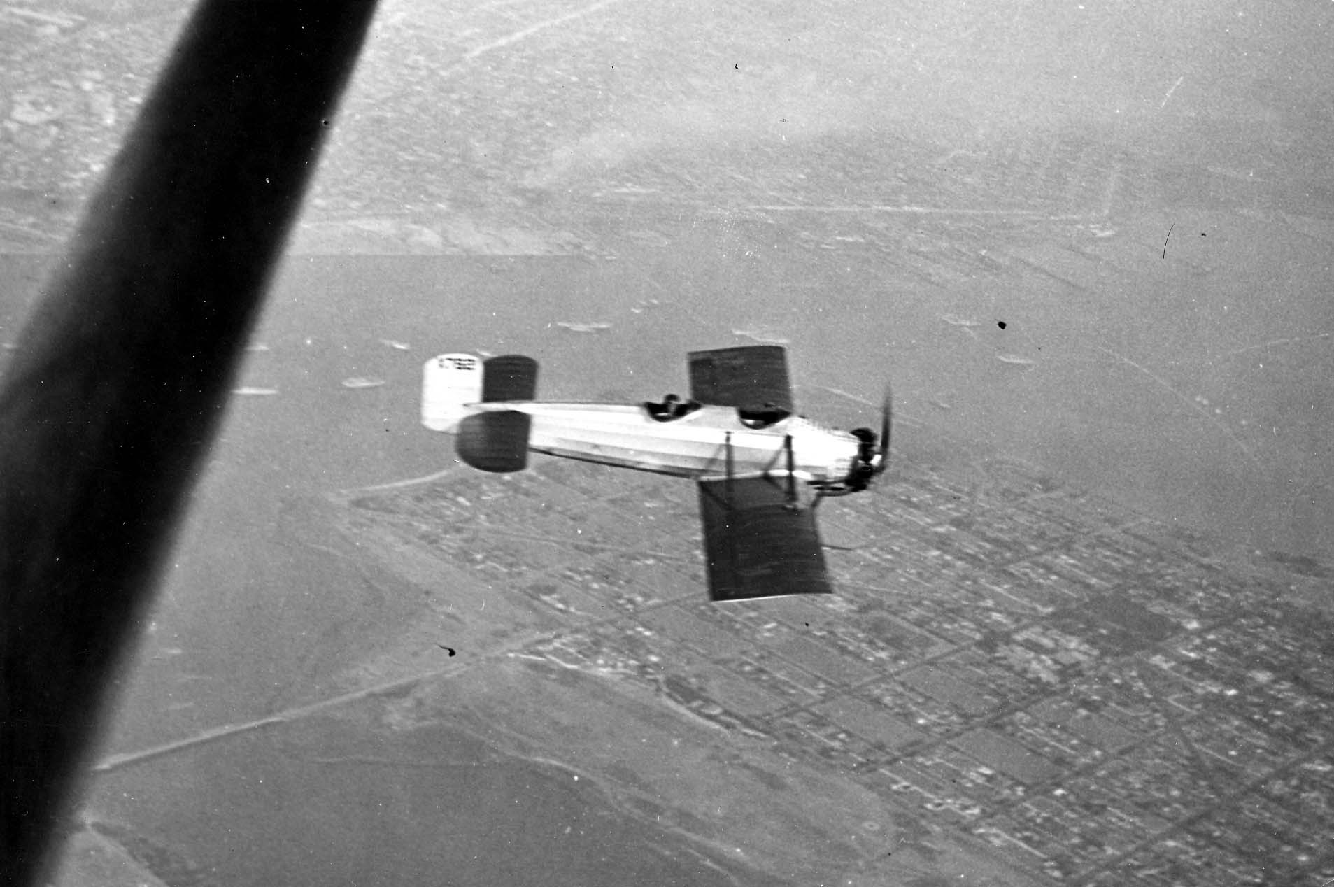 A high-angle air-to-air perspective looking down at the Hall X-1 prototype banking over a vast urban street grid and harbor.