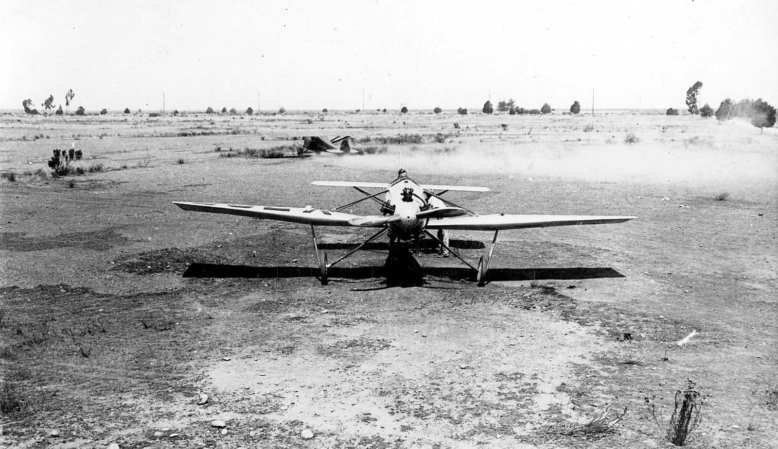 Direct frontal view of the Hall X-1 prototype on a dirt runway with another aircraft taxiing and kicking up dust in the background.
