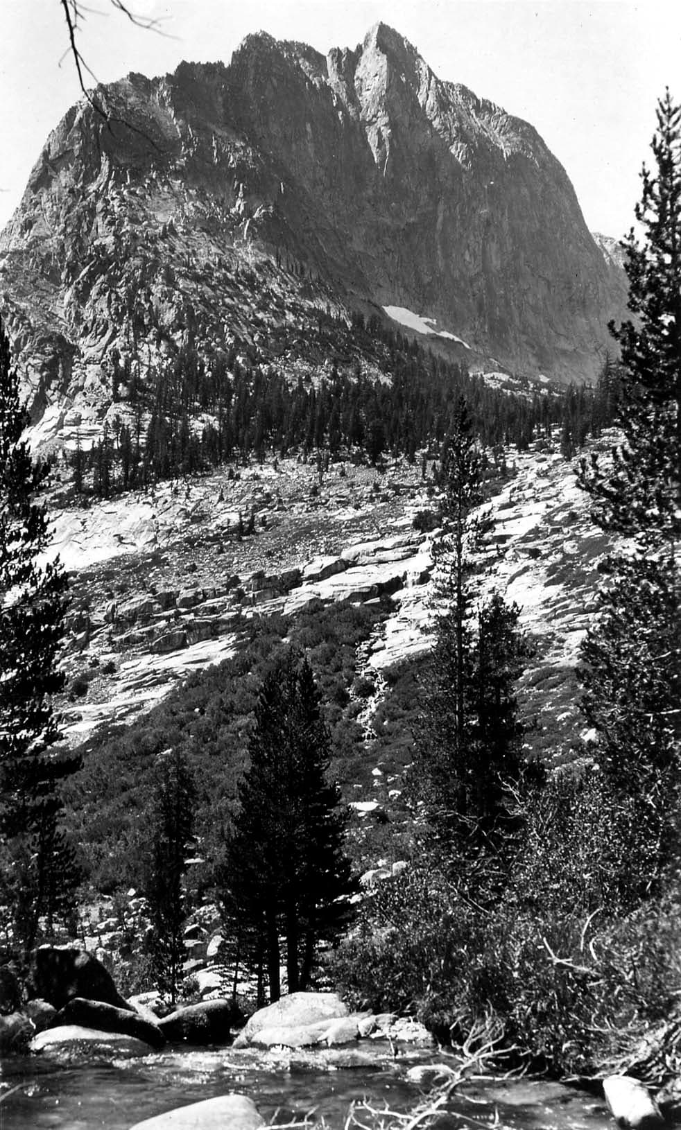 A sheer, dramatic granite peak towering vertically over a boulder-strewn alpine creek and dense pine forest in the Sierra Nevada.