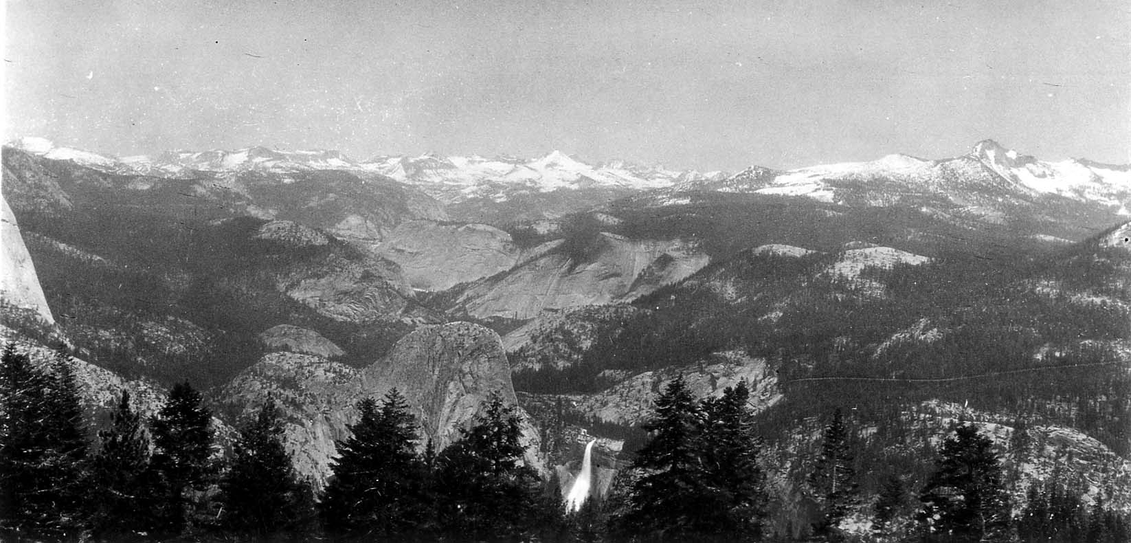 An expansive panorama from Glacier Point showing Liberty Cap, distant Nevada Fall, and snow-capped High Sierra peaks under clouds.