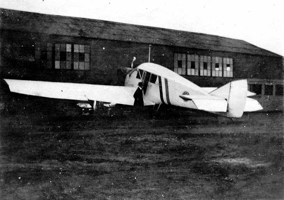 A high-wing commercial cabin monoplane parked on the tarmac outside a large multi-story brick and glass aviation manufacturing hangar.