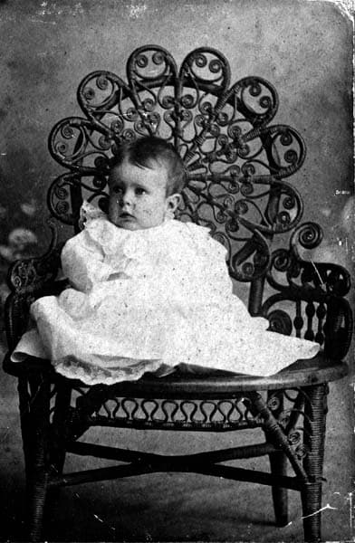Vintage cabinet photograph of Donald A. Hall Sr. as an infant in a white gown seated in a high-backed wicker peacock chair.