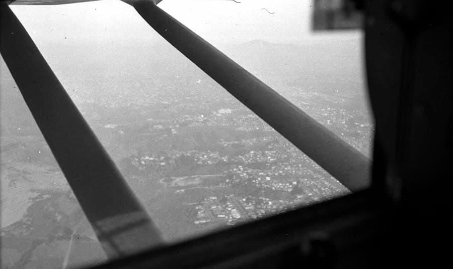 A first-person view looking out the side window of the Spirit of St. Louis in flight, showing the wing struts and the city of San Diego far below.