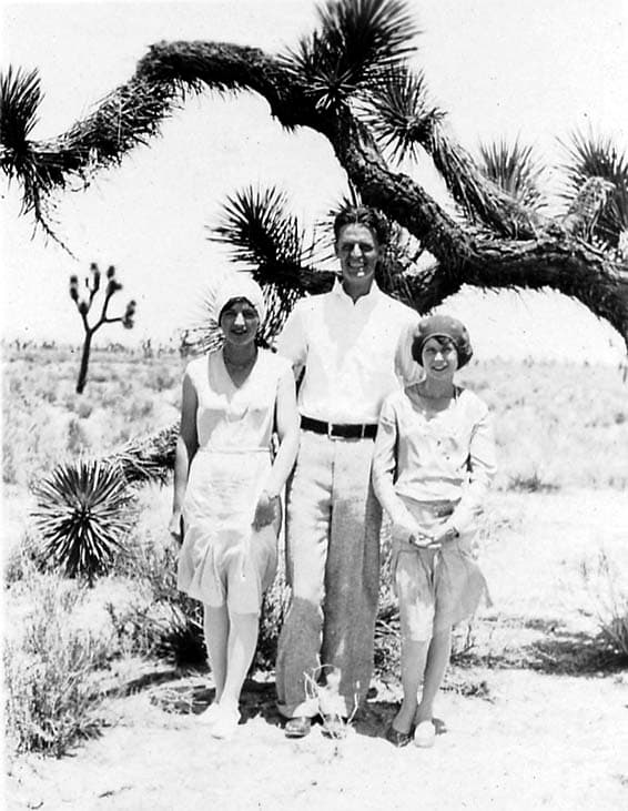 A man and two women in light 1920s summer attire posing together under the twisting limbs of a large Joshua tree in the arid Mojave Desert.