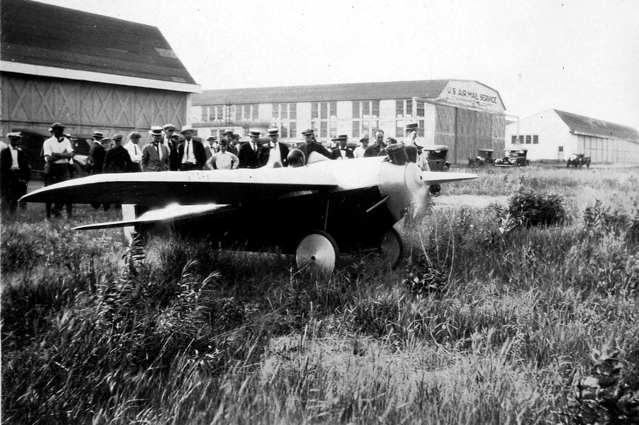 A sleek single-seat monoplane racer with a two-cylinder engine parked in weeds outside a US Air Mail Service hangar before a crowd.