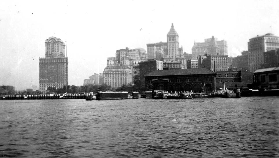 A harbor view of the Lower Manhattan skyline from the water, featuring the arched Pier A fireboat station, the Whitehall Building, and the Standard Oil Building.