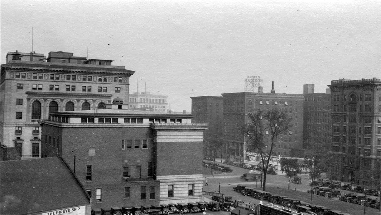 An elevated perspective across downtown Detroit rooftops toward Madison Avenue and the illuminated Hotels Madison-Lenox sign.