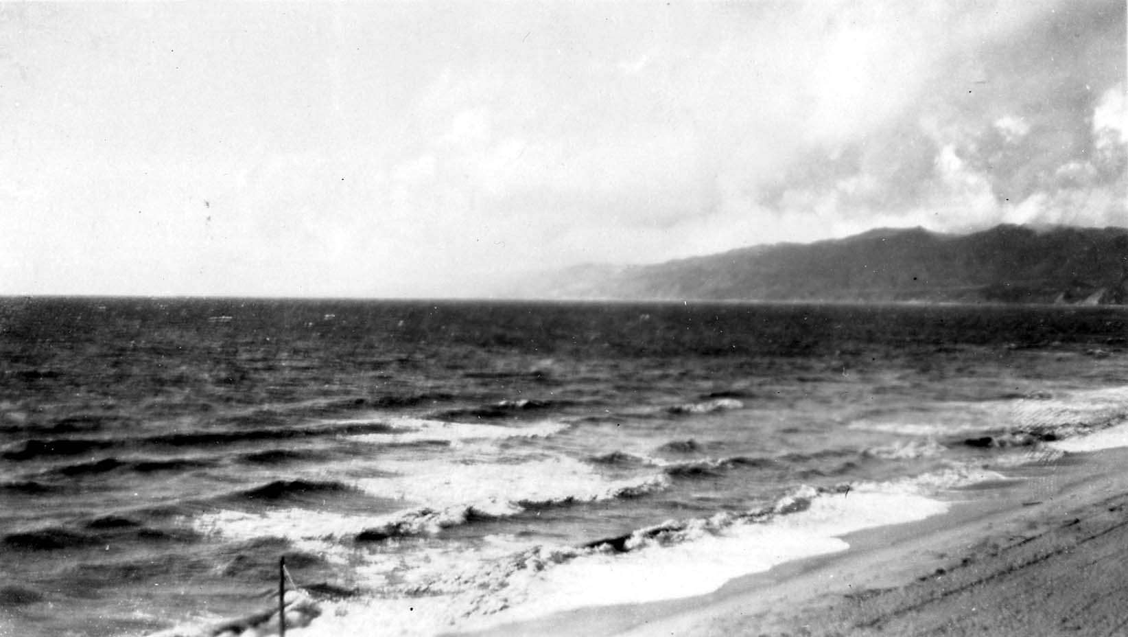 A view along a sandy California beach showing rhythmic ocean breakers washing ashore beneath hazy coastal mountain ridges.