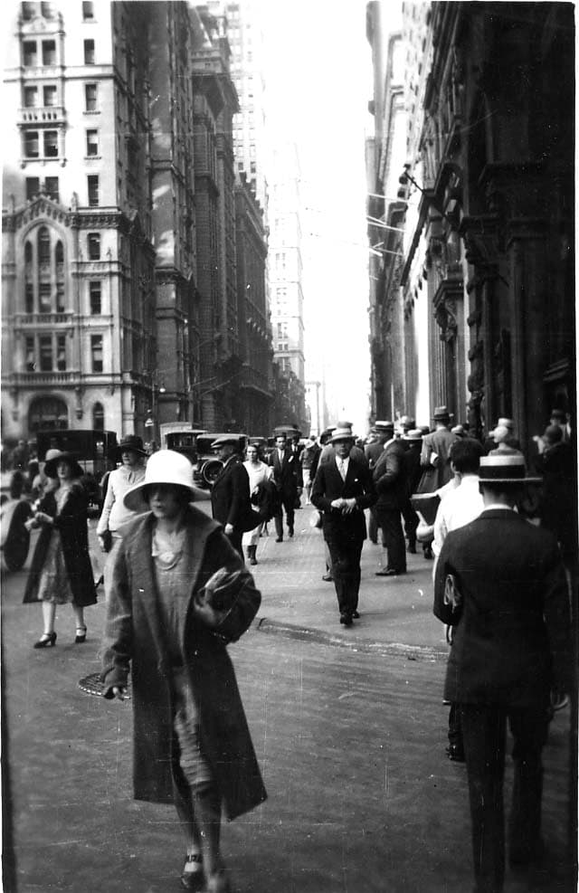 A bustling street-level view looking down a narrow Lower Manhattan canyon filled with pedestrians in 1920s cloche hats and straw boaters during the Lindbergh celebrations.