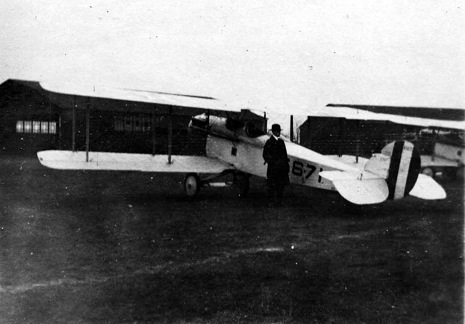 A side profile view of an open-cockpit military biplane trainer bearing fuselage number 671 with an officer in a suit standing by the cockpit.