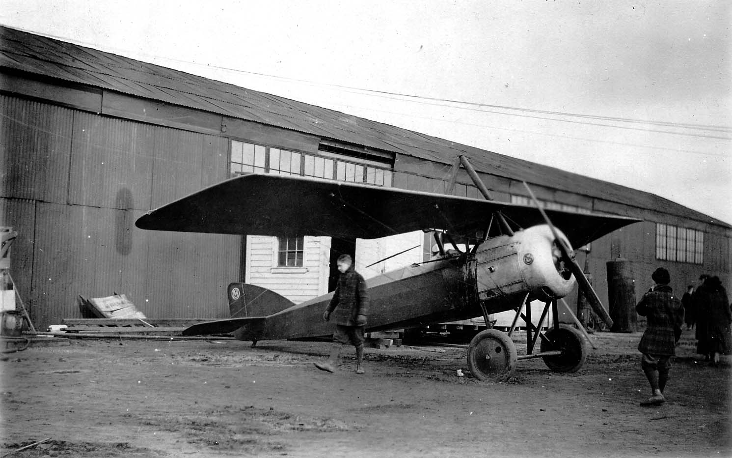 A parasol-wing monoplane with an uncowled rotary or radial engine parked outside a corrugated metal hangar as a young pedestrian walks past.