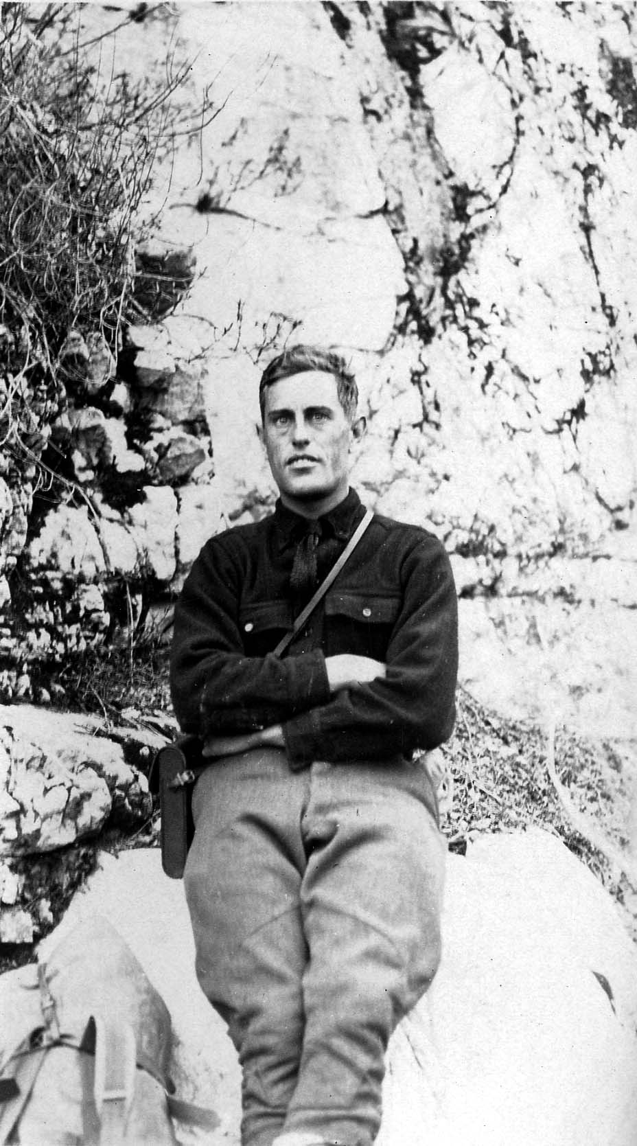 Donald A. Hall Sr. sitting against a sheer granite rock face wearing outdoor breeches, a work shirt, and a leather camera strap.