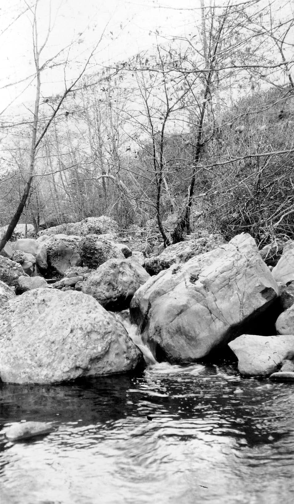 A clear mountain stream flowing between large, weathered granite boulders and bare deciduous trees along the riverbank.