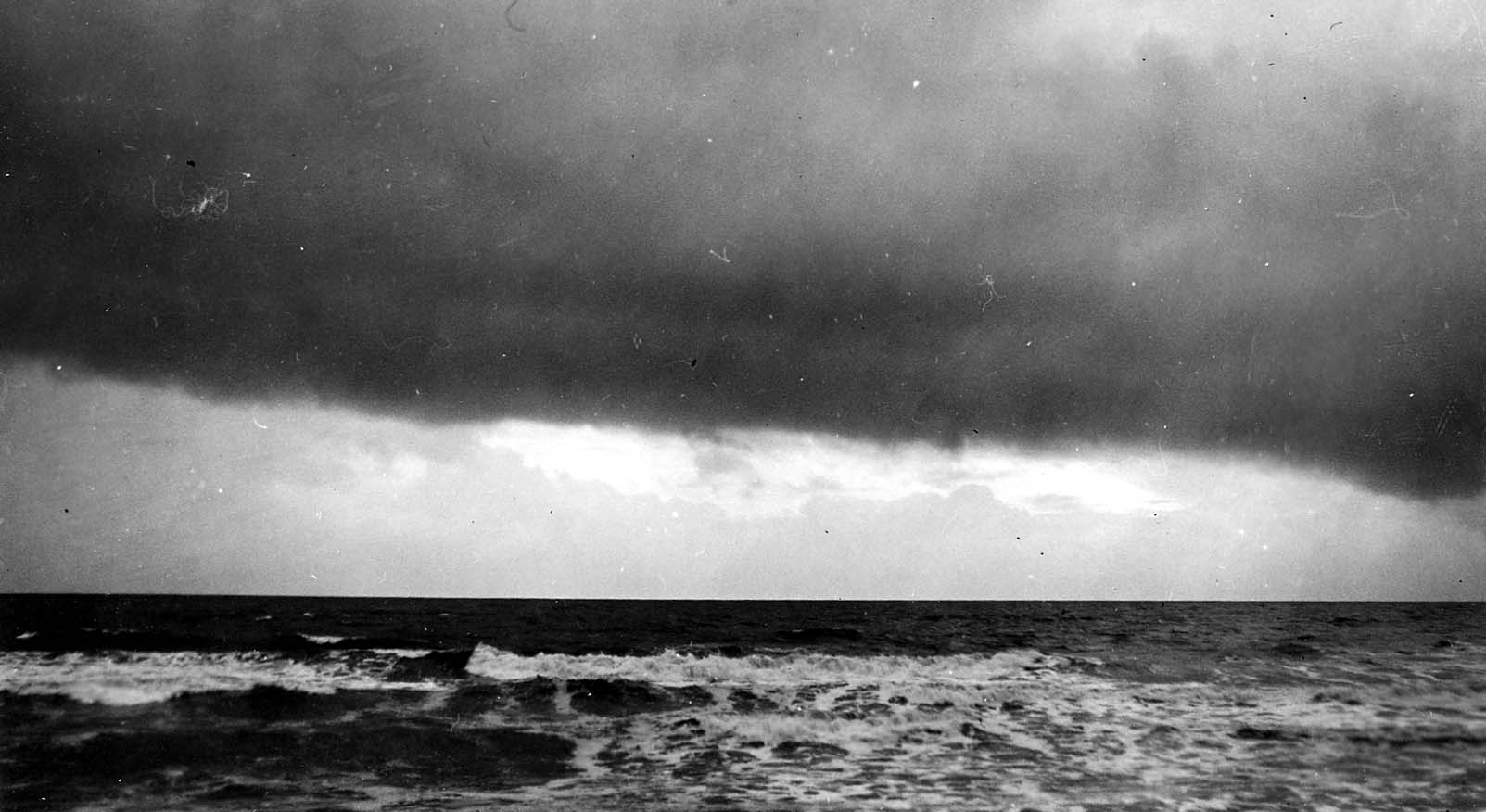 An atmospheric seascape featuring a dark, horizontal storm cloud hanging over the Pacific Ocean horizon above breaking whitecaps.