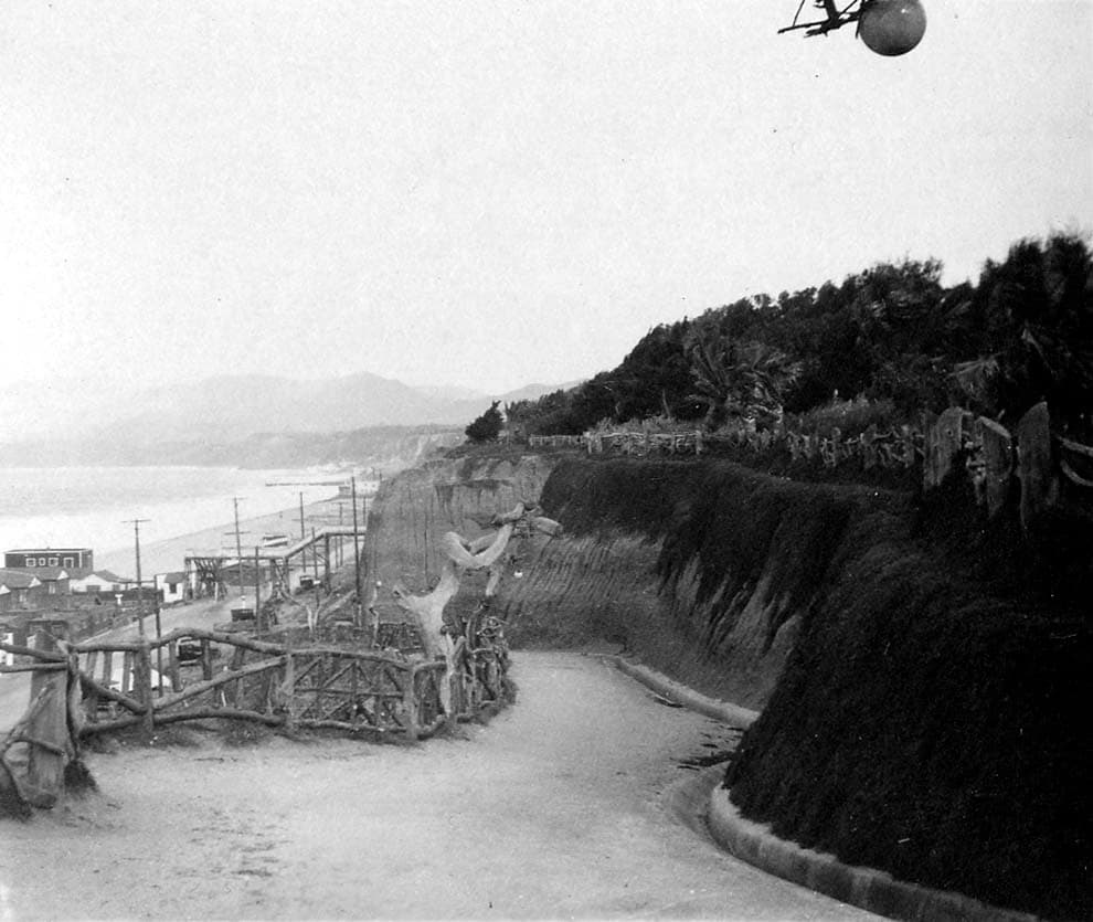 A curving paved pathway in Palisades Park with rustic bentwood fencing, dark hillside foliage, and a decorative spherical light fixture suspended above.