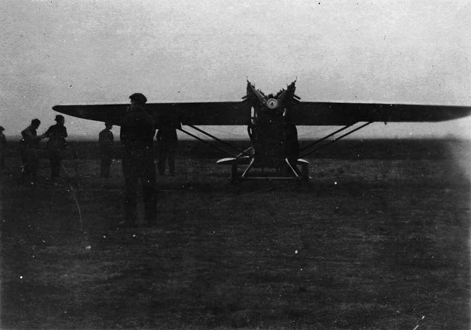 Direct frontal view of an experimental parasol-wing monoplane on an unpaved airfield with spectators standing around the wings.