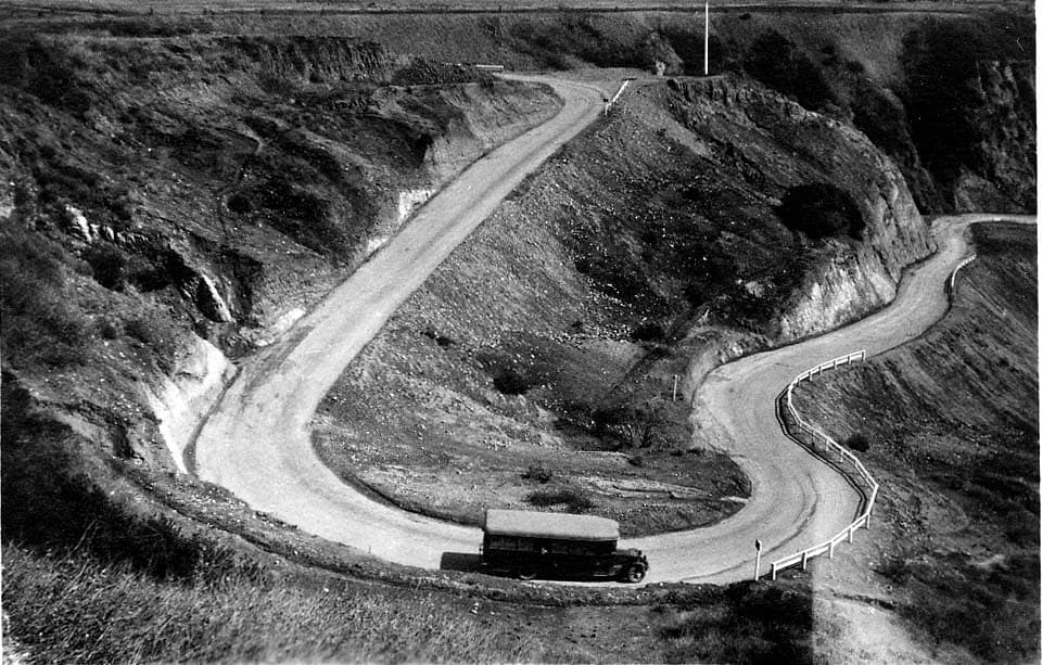 A vintage passenger motorcoach navigating a sharp, unpaved hairpin switchback descending the Santa Monica coastal bluffs.