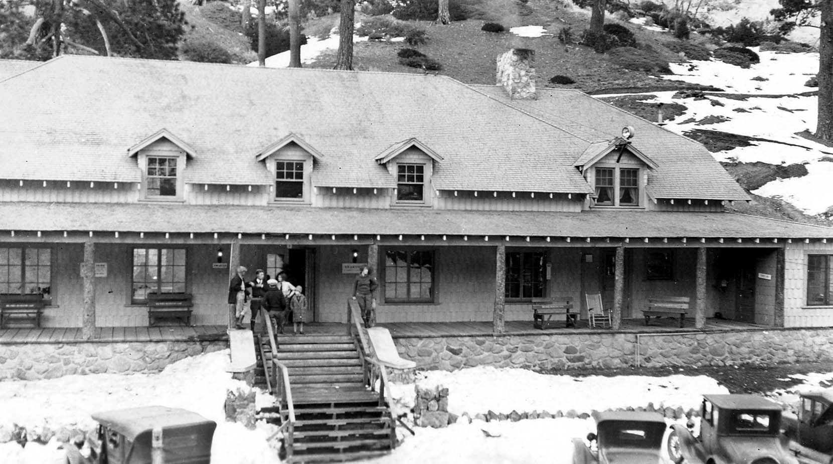 A large rustic mountain lodge with gabled dormers and fieldstone foundation in deep snow with vintage automobiles parked below.