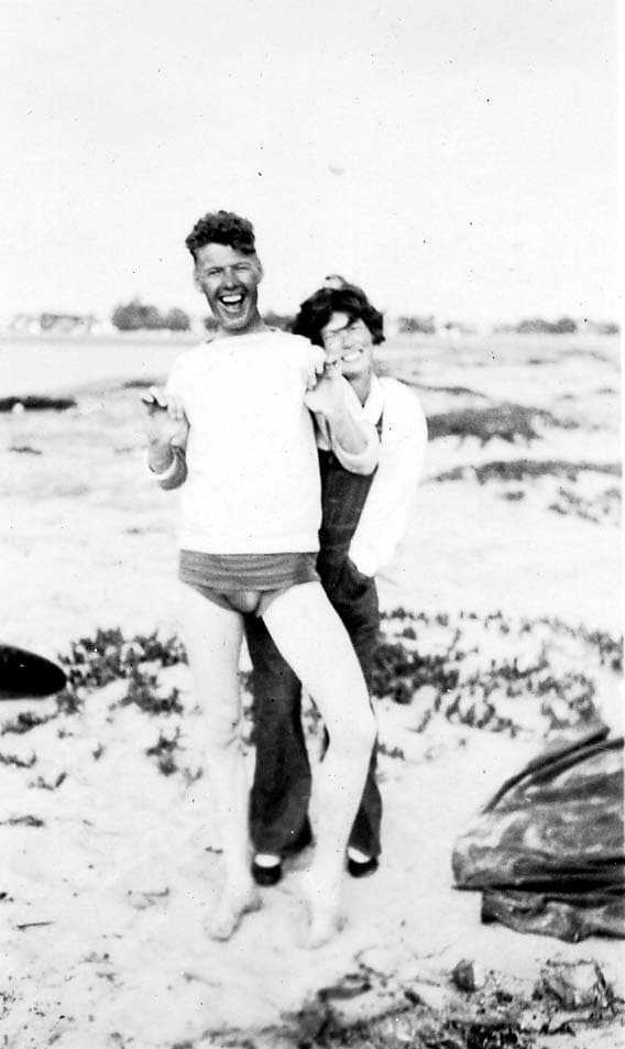 A joyful, candid photograph of a man in swim trunks and sweatshirt laughing on a sandy beach while a woman playfully holds him from behind.