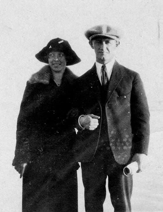A formal portrait of Donald A. Hall Sr. in a tailored three-piece suit and flat cap holding rolled blueprints beside an elegant woman in a fur-collared coat and cloche hat.