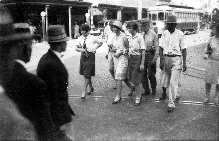 Pedestrians in 1920s fashion, including women in cloche hats and men in work shirts, crossing a wide city street in front of an electric streetcar.