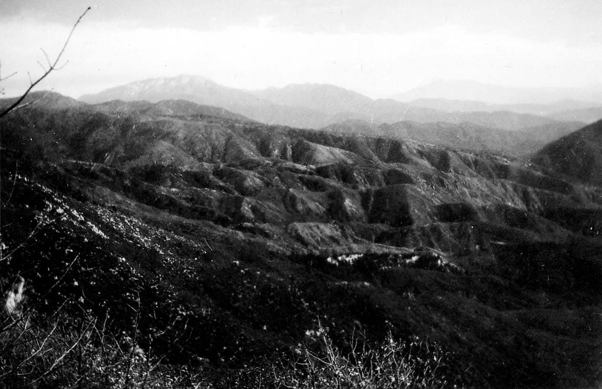 A high-altitude panoramic view of rolling, heavily creased mountain ridges receding into distant hazy peaks under an overcast sky.