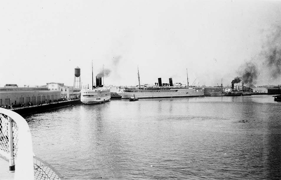 A harbor view from a vessel showing the Los Angeles Steamship Company terminal warehouse, water tower, and the twin-funneled passenger liner SS Yale or SS Harvard.