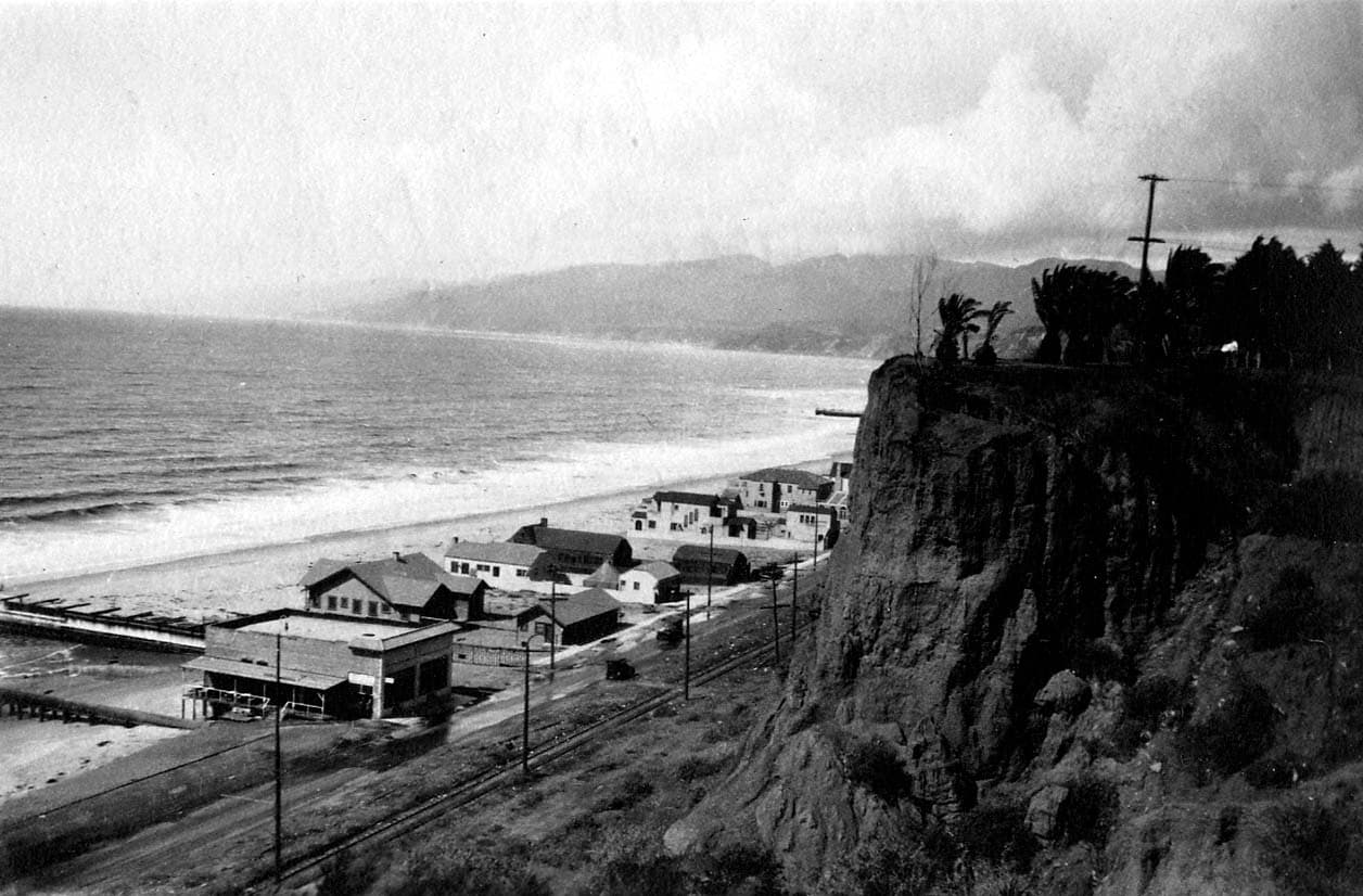 A steep vertical perspective looking down a sheer Palisades cliff face to coastal railroad tracks, beach bungalows, and the Pacific Ocean.
