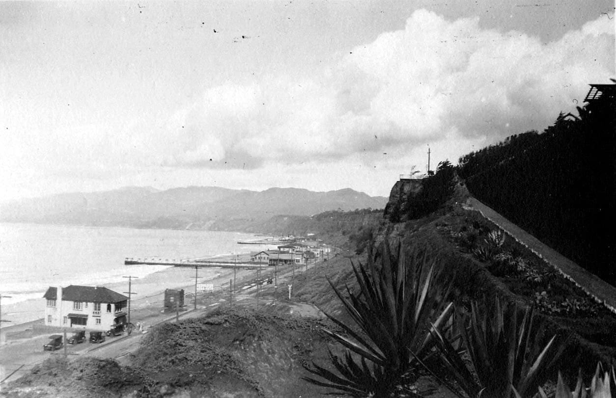 A high-angle elevated view from the Santa Monica Palisades looking northwest along the beach, coastal highway, and tracks toward the distant Malibu mountains.
