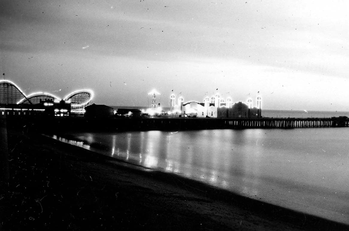 An evening view across the beach toward the illuminated Santa Monica Pier, showcasing the Moorish arches of the La Monica Ballroom and the adjacent roller coaster.