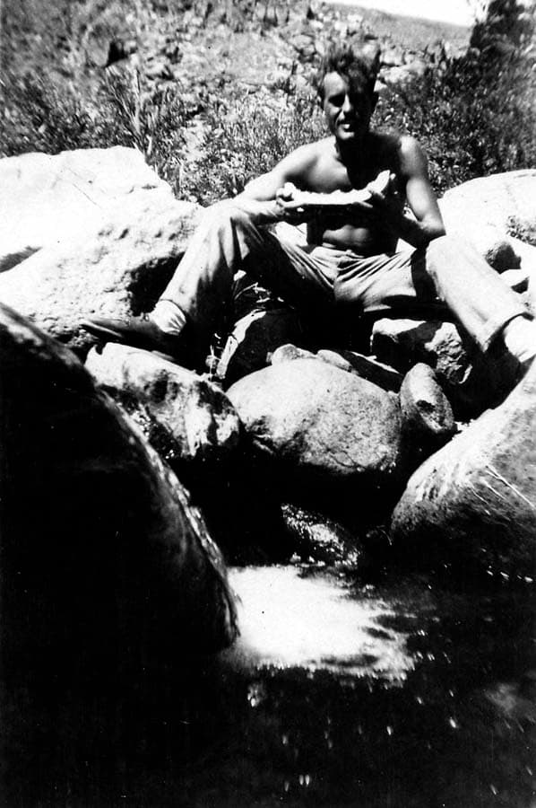 Outdoor photograph of Donald A. Hall Sr. sitting shirtless on mountain boulders eating watermelon beside a stream.