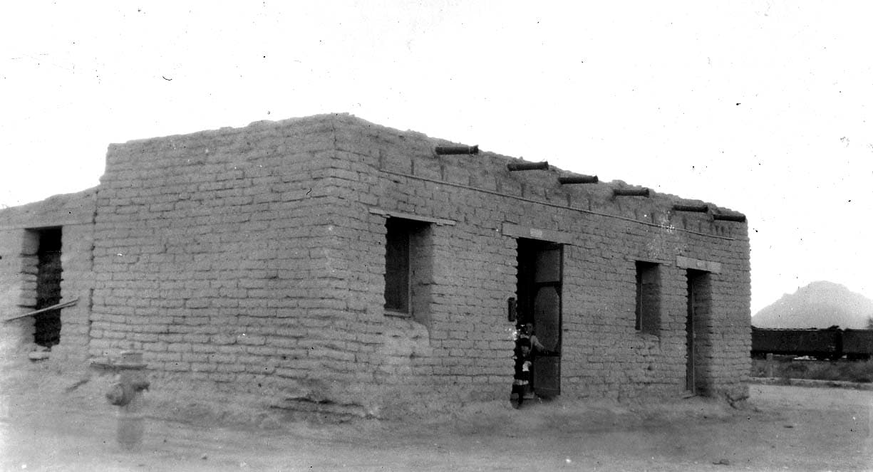 A weathered flat-roofed mud-brick adobe building with projecting log vigas and a young child in the doorway beside railroad tracks and a desert butte.