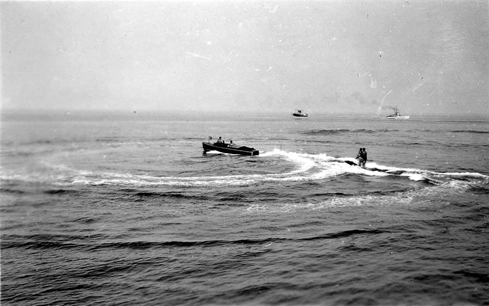 A wooden runabout speedboat carving a turn while towing an aquaplaner standing on a surf board, with a float biplane and ship in the distant background.