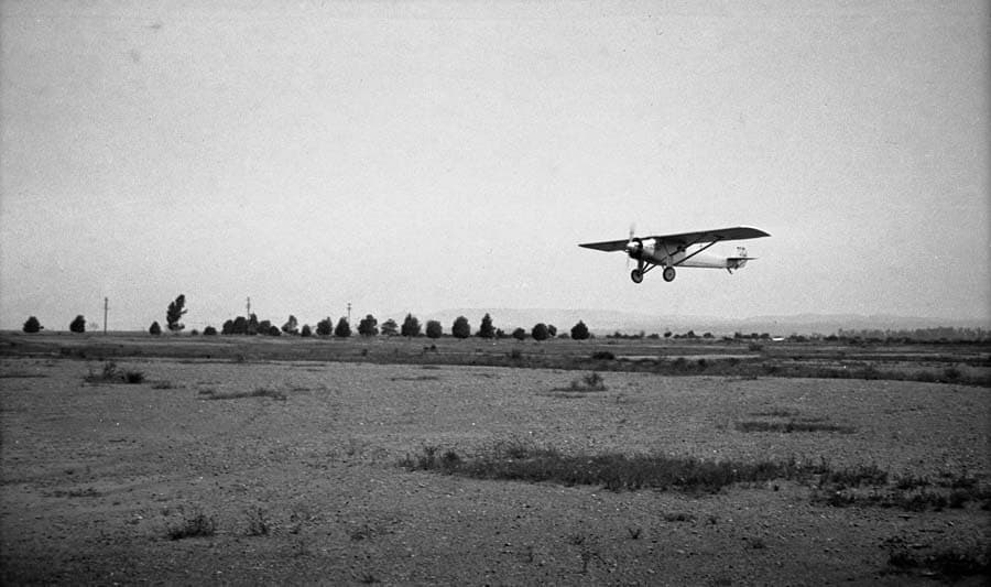 An air-to-air photograph of the Spirit of St. Louis flying low over the flat, brush-covered terrain of the Camp Kearny parade grounds.